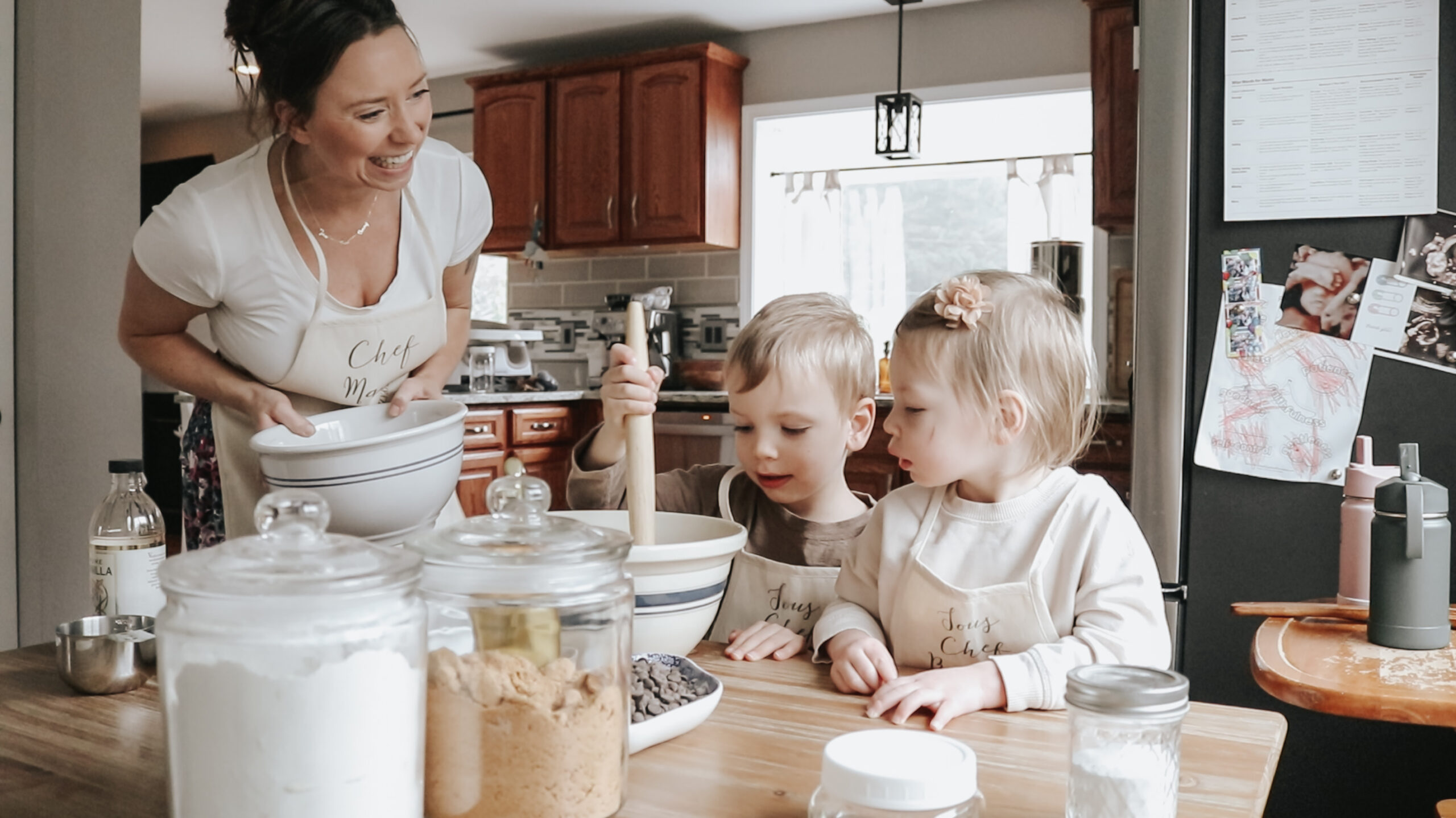 Christian homemaker sttanding at a table with her children holding a bowl while her two children help bake, one child is stirring and the other is watching